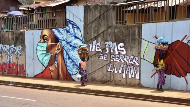 Des femmes passent devant un graffiti sur un mur illustrant les mesures d'hygiène visant à freiner la propagation du coronavirus COVID-19 à Conakry, en Guinée.