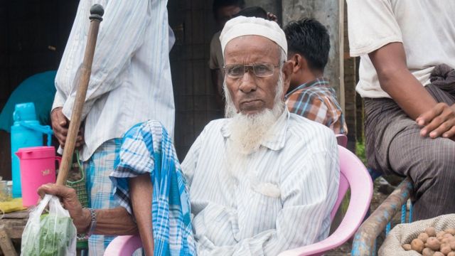 An older Muslim man with a white beard and glasses under his cap sits at the market in Maungdaw, holding a walking cane by his side