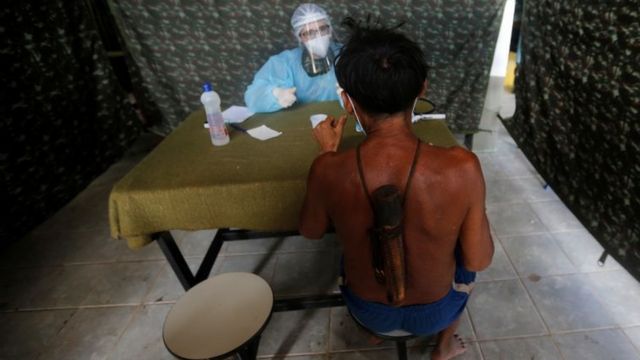 Em tenda de atendimento, homem indígena de costas é atendido, do outro lado da mesa, por profissional de saúde todo coberto por jaleco e face shield