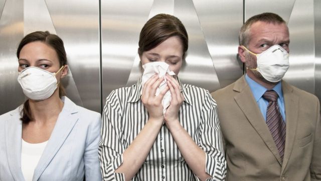 Três pessoas no elevador, uma assoando o nariz e outras duas de máscara