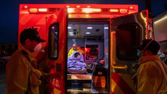 Dois paramédicos fecham as portas de uma ambulância, onde um terceiro paramédico atende um paciente encamado
