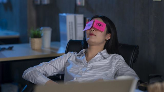 A woman asleep in an office chair with post-it notes over her eyes