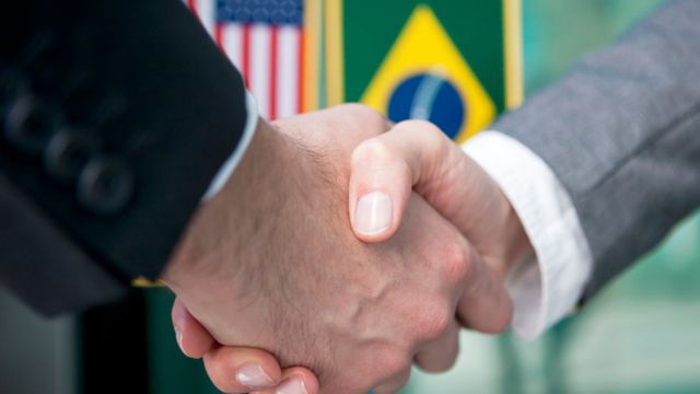 Duas pessoas se cumprimentam com aperto de mão, com bandeiras do Brasil e EUA ao fundo
