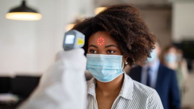 A woman wearing a surgical mask has her fever measured with a reader on her forehead