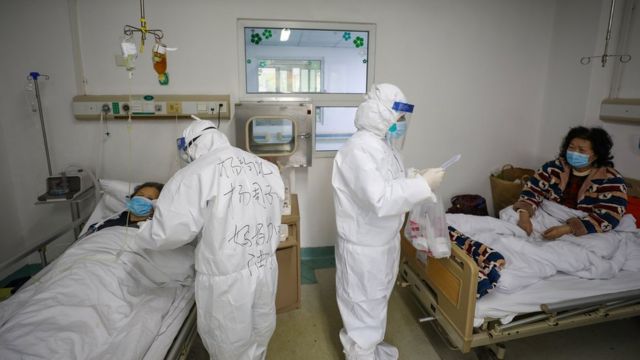 Dois profissionais de saúde, cobertos por máscaras e jalecos, falam com duas pacientes deitadas em maca
