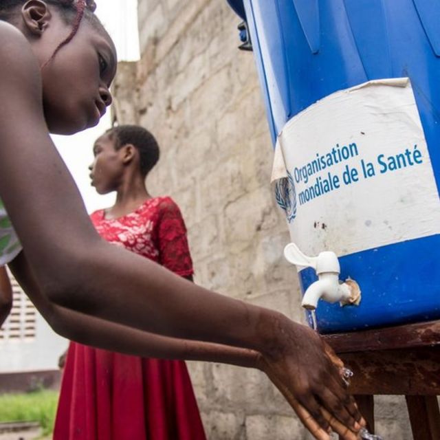 Una niña se lava las manos con agua de un bote que dice "Organizacón Mundial de la Salud".