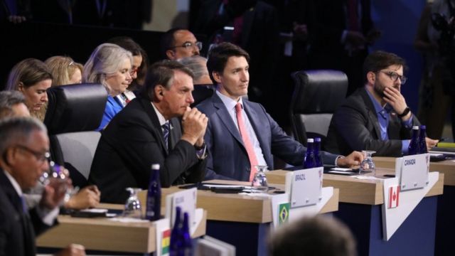 O presidente Bolsonaro ao lado do primeiro-ministro do Canadá, Justin Trudeau, sentados na plateia e com outras pessoas em volta