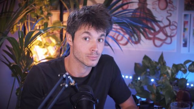 Image d'un YouTuber assis dans une pièce faiblement éclairée, avec un micro devant lui et des plantes en pot derrière lui. Il regarde droit vers la caméra et a les sourcils relevés.