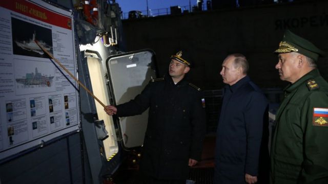 Photograph shows two white men in military uniform pointing a ship on an information board for President Vladimir Putin with a dark background