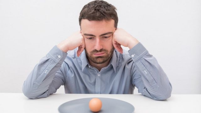 Hombre joven mirando a un plato con un huevo con cáscara.