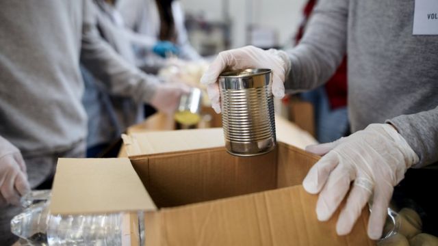 Duas pessoas em frente a caixa de papelão apoiada em mesa com comida dentro; uma das pessoas coloca uma lata na caixa