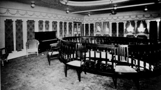 Foto em preto e branco mostra uma sala com cadeiras e um piano
