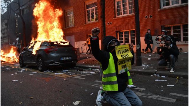 A French protester kneeling on the ground and raising his fist in the air, next to a burning car
