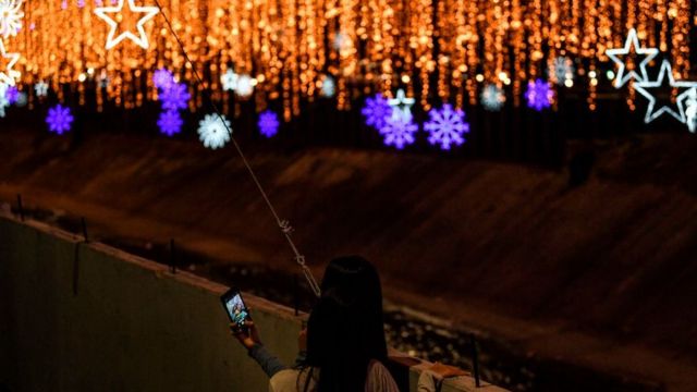 Em foto noturna, mulheres tiram selfie na margem de rio em Caracas, rodeado por luzes de Natal