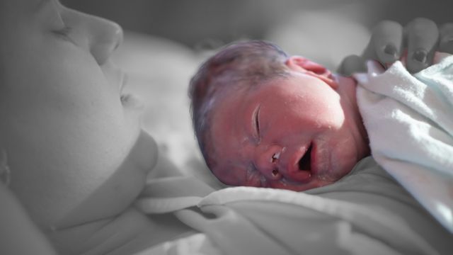 Deitada, mulher segura um bebê recém-nascido no colo; o bebê possui uma camada branca de vérnix caseoso sobre a pele