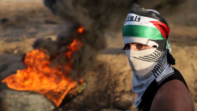 A masked Palestinian protester passes by burning tyres during clashes with Israeli troops near the border between Israel and Central Gaza Strip October 14, 2015.