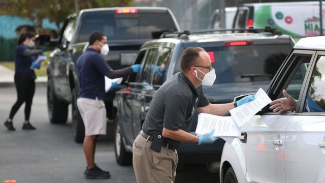 Personas con mascarillas entregando aplicaciones para la solicitud de desempleo a automovilistas en Hialeah, Florida.