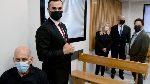 إيهود أولمرت (من اليسار) يجلس داخل محكمة في تل أبيب، بينما يقف خلفه بنيامين نتنياهو (الثاني من اليمين) وزوجته سارة (10 يناير/كانون الثاني 2022)