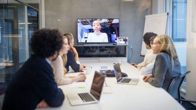Reunión en Zoom con una participante en una pantalla y el resto de un equipo reunido en una mesa.