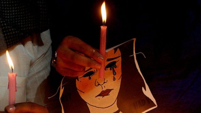 Em foto noturna, mão segura cartaz com desenho de mulher chorando e uma vela