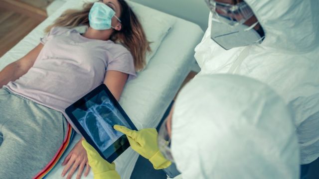 Dois profissionais de saúde paramentados e com jaleco olham para tablet, que mostra uma imagem de raio x; atrás, uma mulher jovem de máscara deitada em maca