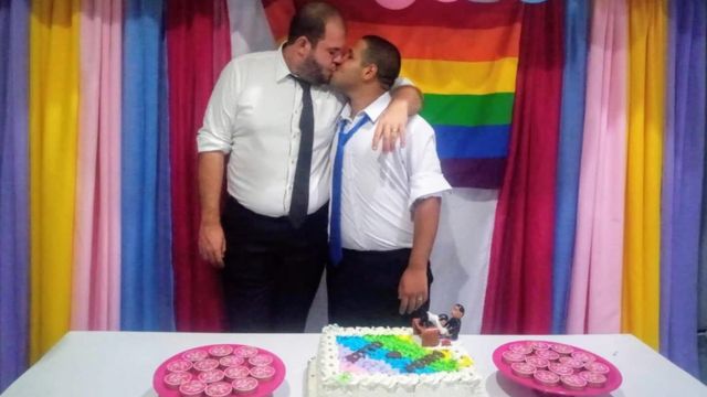Pedro e o marido se beijam em casamento com bolo e bandeira LGBT ao fundo