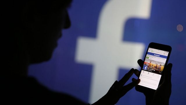 Homem na contraluz mexe no aplicativo do Facebook no celular, com grande logo da rede social projetada ao fundo