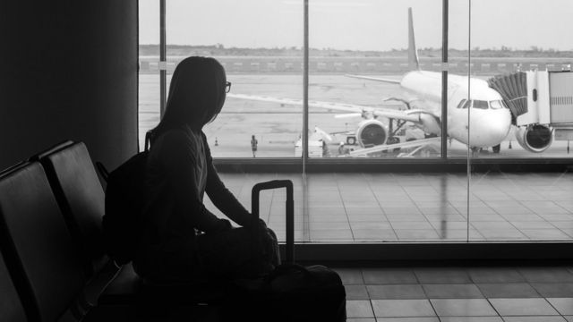 Foto de banco de imagens com jovem segurando uma mala e esperando para embarcar em um saguão de aeroporto, olhando para o avião pela janela