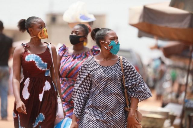 Women wear face masks with kente and wax print designs as they walk through a market.