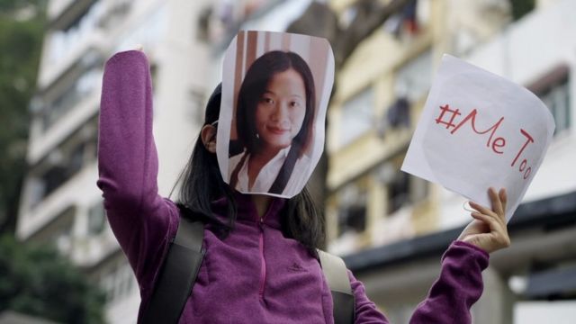 Seorang pengunjuk rasa memegang spanduk MeToo dan mengenakan topeng wajah Huang