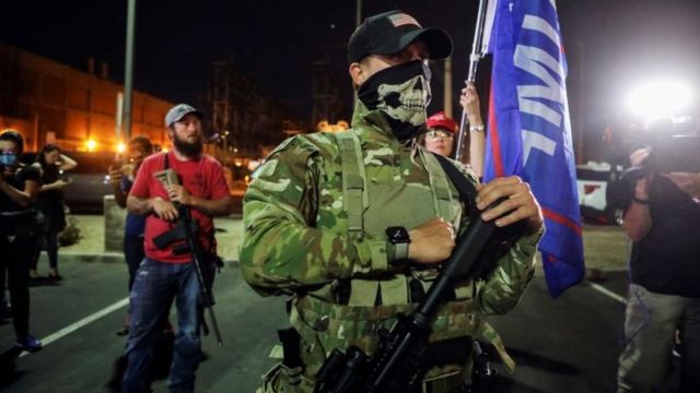 Em foto noturna, homem porta fuzil; atrás, outro homem armado e bandeiras com nome de Trump