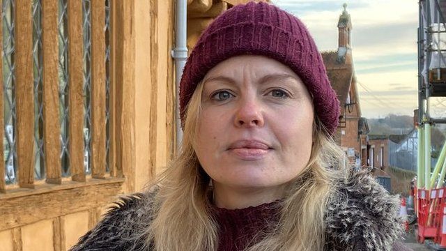 Bernadette Ashcroft wearing a coat and a Beanie hat outside one of the Tudor buildings, typical for the scenery in Lavenham.