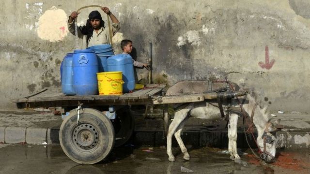 Um homem enche recipientes de plástico com água em um carrinho puxado por um burro