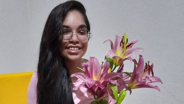 Rute atualmente, com 16 anos, segurando uma flor e sorrindo para a foto. Ela tem cabelos longos pretos e usa óculos de grau