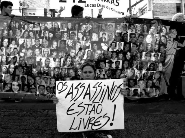 Mulher em frente a mural com fotos das vítimas segurando cartaz em que se lê: 'Os assassinos estão livres'