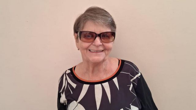 Sônia, 67, sorri em frente a uma parede beige; sua pele é branca, ela tem cabelos grisalhos e curtos e usa óculos escuros, uma blusa 