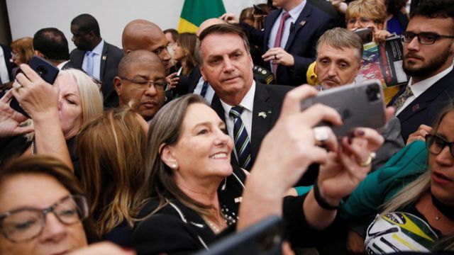Bolsonaro sorri para selfie, rodeado de apoiadores em ambiente fechado
