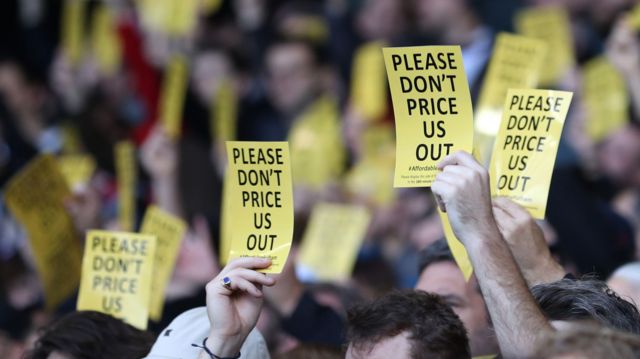 Fulham fans hold cards saying "Please Don't Price Us Out" at a Premier League match