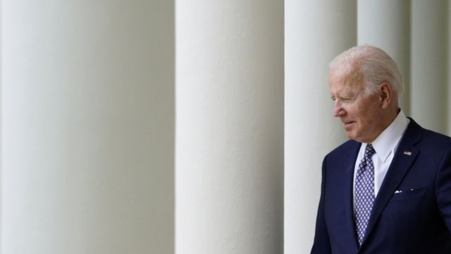 Biden caminhando na Casa Branca, sorrindo timidamente