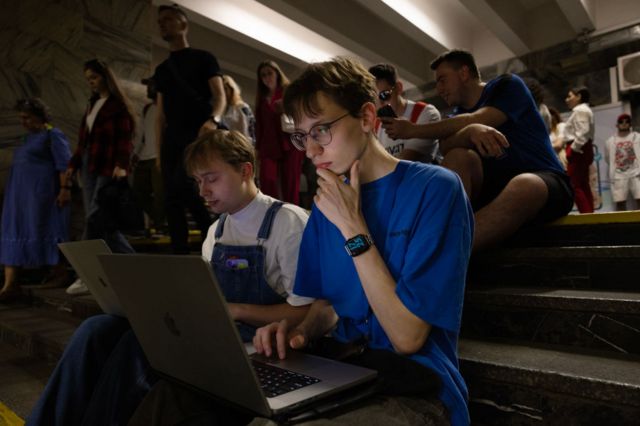 Dos jóvenes trabajando en sus laptops dentro de un refugio antiaéreo.