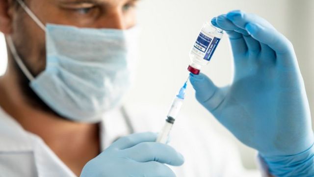 Profissional de saúde segura uma ampola e, com uma seringa, extrai a vacina tríplice viral