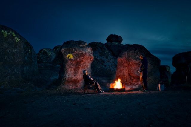 A couple sit in the darkness illuminated by a small camp fire