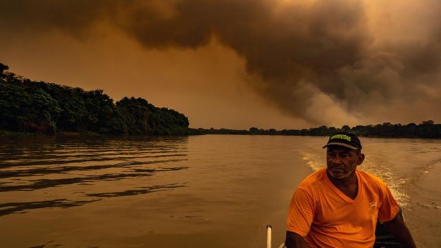 Homem em um barco com fumaça de grandes proporções ao fundo