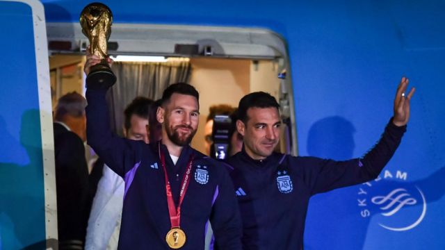 Lionel Messi sostiene la Copa FIFA en la puerta de una avión, acompañado del director técnico de la selección argentina de fútbol, Lionel Scaloni.