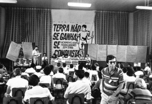 Sem terra reunidos em salão, com cartaz ao fundo com o lema "terra não se ganha, se conquista"