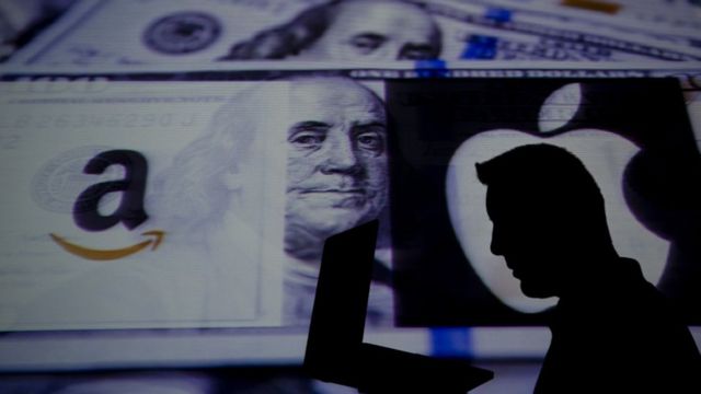 Sombra de homem em frente a tela com imagem de nota de dólar e logos da Amazon e Apple