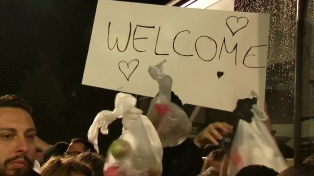 Someone waving a sign saying "Welcome" with bags of apples and water for migrants arriving at Saalfeld station in east Germany