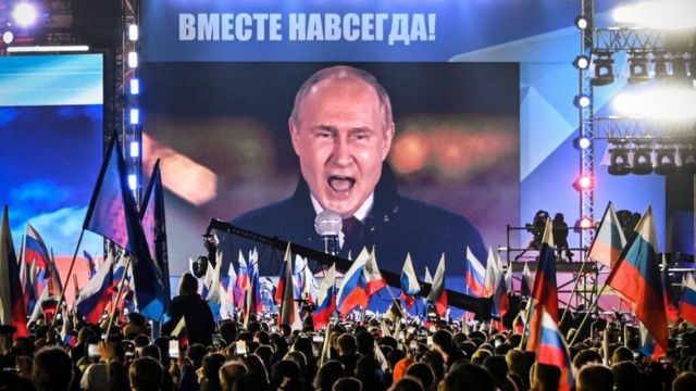 Vladimir Putin fala para multidão em Moscou, com as palavras "Juntos para sempre" no topo da tela.