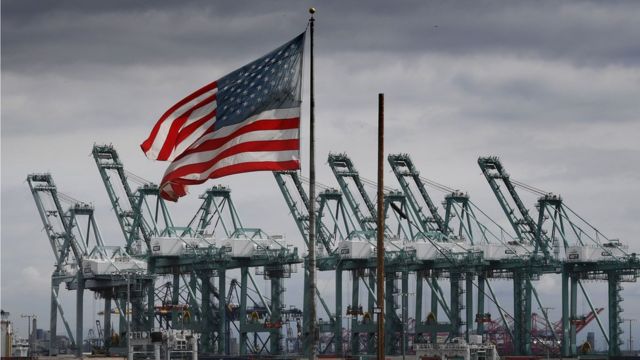 The US flag flies over shipping cranes and containers in Long Beach, California on March 4, 2019.
