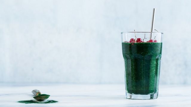 Un grand verre de smoothie aux fruits verts avec de la spiruline hawaïenne et des groseilles rouges sur le dessus. Vu sur une table en marbre blanc, et une cuillère de spiruline à côté.